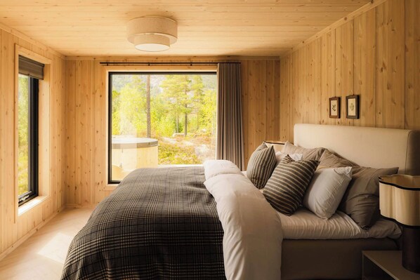 A wooden-paneled bedroom with a large bed, a bedside table, and a window with a view of trees.