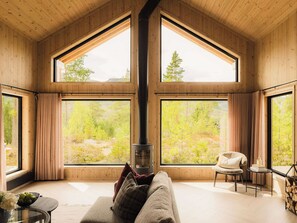 A cozy room with a large fireplace, wooden walls, and large windows offering a view of a forest.