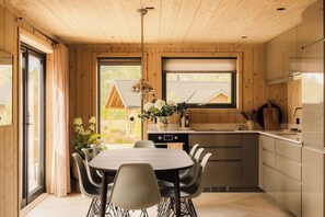 A modern kitchen with a dining area, wooden walls, and a view of a house through the window.