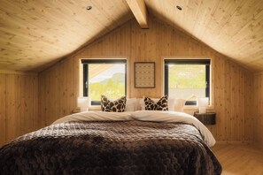 A bedroom with a wooden ceiling, a large bed with a patterned bedspread, two windows with a view of greenery, and a framed picture on the wall.