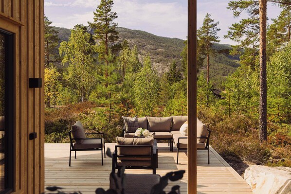 An outdoor deck with a sofa and chairs, overlooking a forested landscape with mountains in the distance.