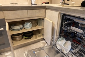 A kitchen with a dishwasher, a sink, and a cabinet with stacked plates and bowls. - 2-Bedroom Apartment in Tranquil Tbilisi Perfect for Relaxing Stays (T'bilisi)
