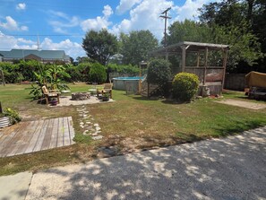 Ein Garten mit einer Holzterrasse, einer Feuerstelle, Gartenmöbeln, einem überdachten Sitzbereich und einem Swimmingpool.