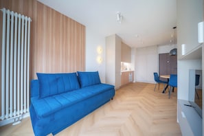 Un espai de vida modern amb un sofà blau, un mur de panells de fusta i una zona de cuina amb una taula de dinar i caires.