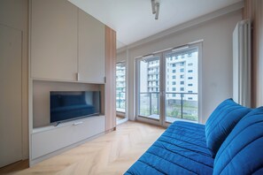 A modern living room with a flat-screen TV, a blue sofa, and large windows offering a view of a building outside. -  Kierbedzia Blue Sapphire Residence (Warszawa)