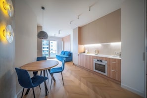 A modern kitchen with wooden cabinets, a built-in oven, and a dining area with a round table and blue chairs. -  Kierbedzia Blue Sapphire Residence (Warszawa)