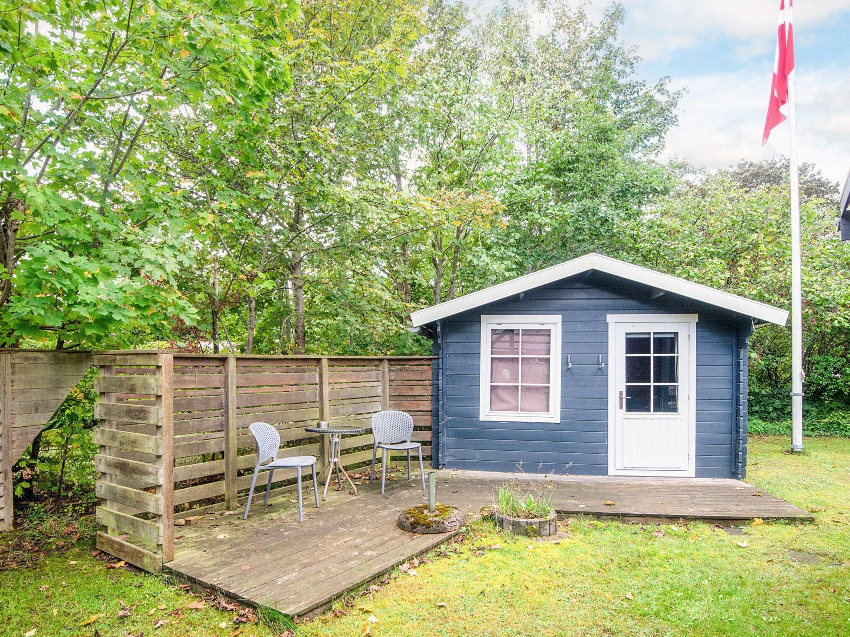 Eine kleine, blaue Holzhütte mit einer weißen Tür und Fenstern, eine Holzterrasse mit zwei Stühlen und einer Fahnenstange mit einer Wimpel.