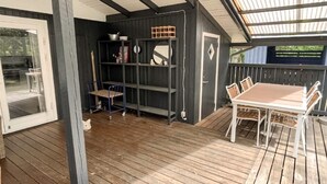 Eine überdachte Terrasse mit Holztisch und -stühlen, einem Metallregal und einer Glastür, die zu einem Außenbereich führt.