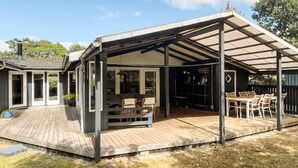 Eine überdachte Terrasse im Freien mit Holzdeck, einem Essbereich mit Stühlen und einer schwarzen Wand mit Regalen.