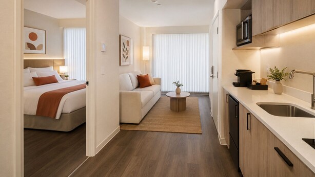 A modern hotel room with a bed, a sofa, a small round table, and a kitchen area with a sink and microwave. - District Liora Hotel (Toronto)