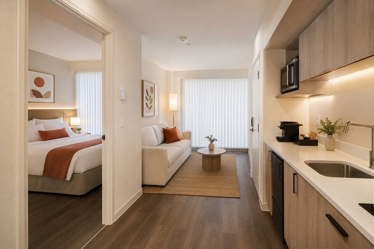 Een moderne hotelkamer met een bed, een slaapbank, een klein rond tafeltje en een kitchenette met een spoelbak en magnetron.