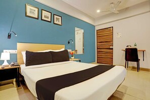 A hotel room with a large bed, a nightstand with a lamp, a desk with a chair, and a wooden door. - Classic Bhumi Stay inn (Bhiwandi)