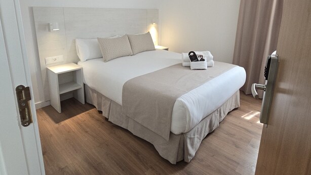 Quarto de hotel com cama, mesas de cabeceira, luminária fixada na parede e porta com puxador.