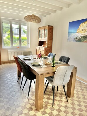 A dining room with a wooden table set for four, a large window with a view of greenery, and a painting of a cliff on the wall.