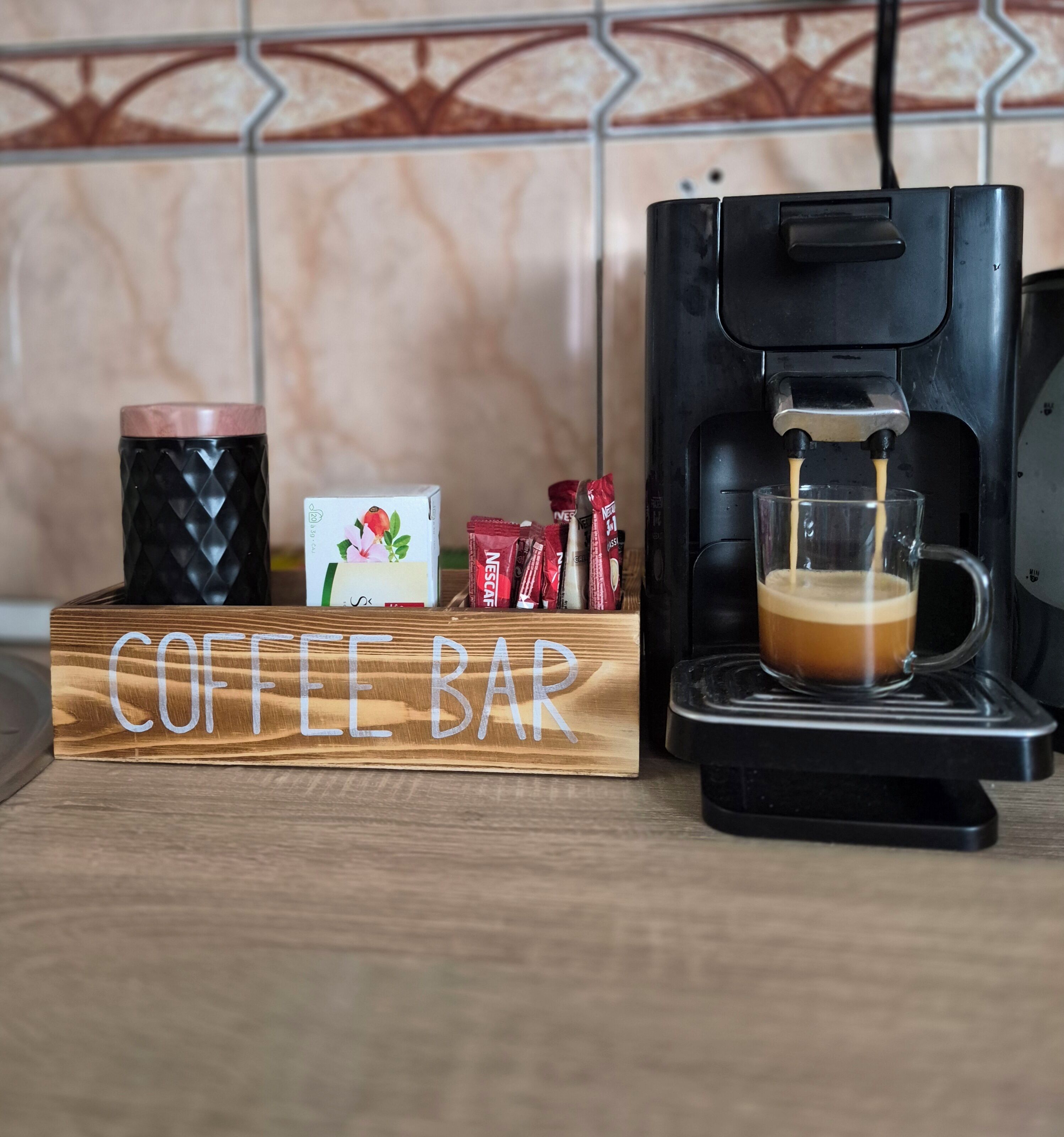 Un coin café aménagé avec une machine à café, une tasse en verre et des dosettes de café, le tout disposé sur une planche en bois portant l’inscription « BAR À CAFÉ ».