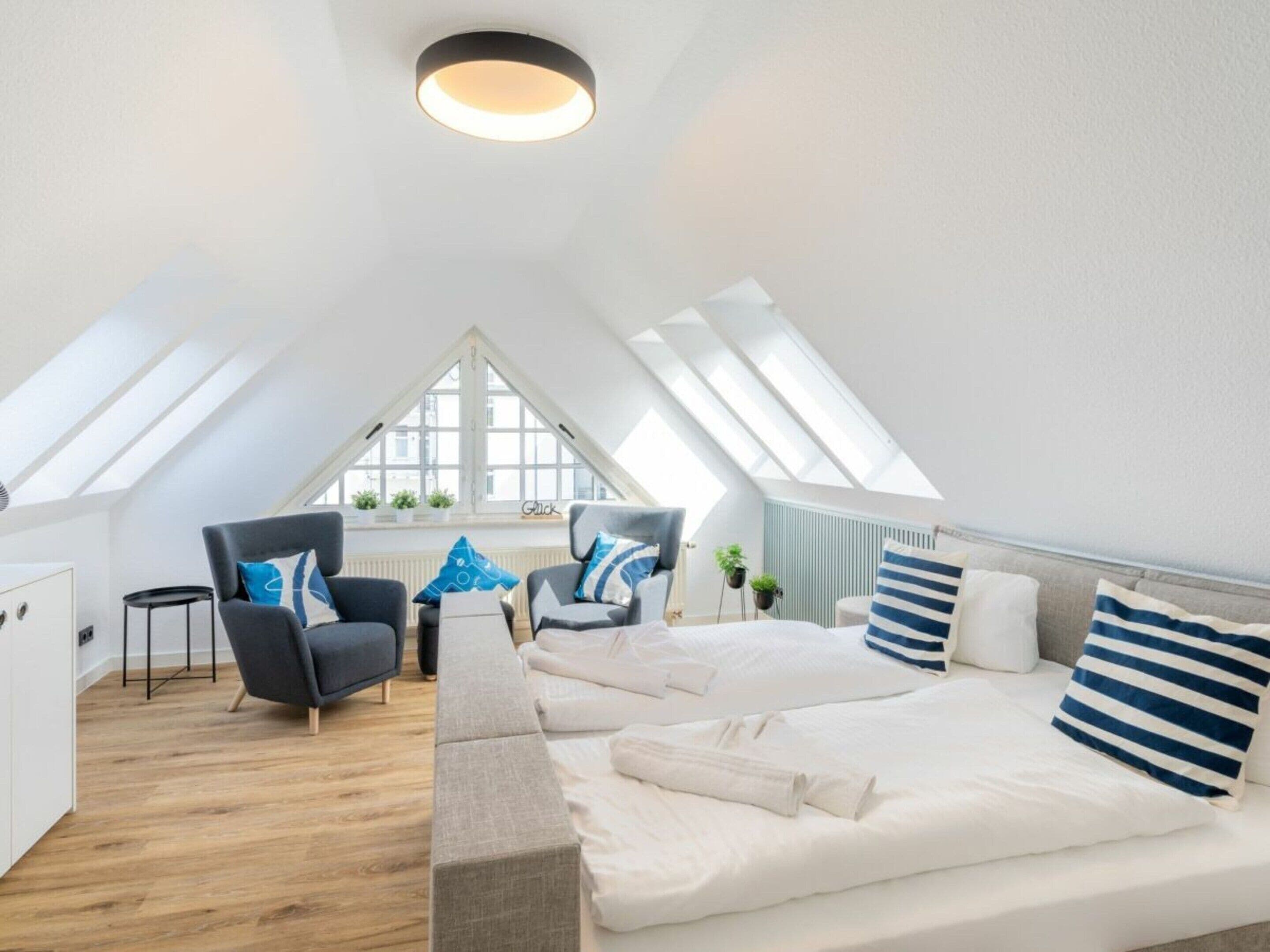 Et moderne loftværelse med skråvæg, ovenlysvinduer, en stor seng, to lænestole og et lille bord.