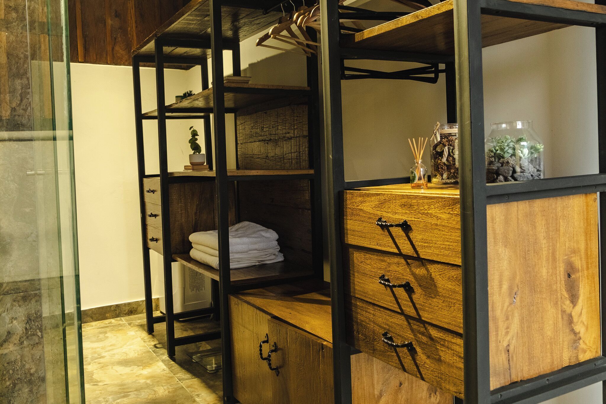 Una habitación con un mueble de madera con cajones, un estante con toallas dobladas y un perchero con ropa.