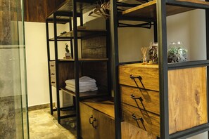 Una habitación con un mueble de madera con cajones, un estante con toallas dobladas y un perchero con ropa.