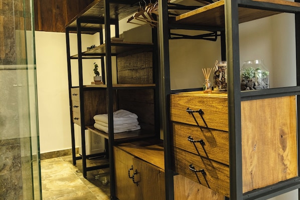 A room with a wooden cabinet with drawers, a shelf with folded towels, and a hanging rack with clothes.