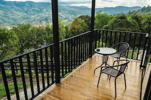 Una terraza de madera con barandilla negra, una mesa redonda y dos sillas de mimbre, con vistas a un paisaje montañoso.