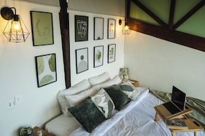 Un dormitorio con una cama, almohadas, una laptop sobre una mesita de noche de madera y una obra de arte colgada en la pared.