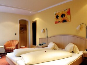 A hotel room with a double bed, a chair, a nightstand, and a painting of sunflowers on the wall. - Shared Rooms in the Main Building - Landhotel Kaserer (Bramberg am Wildkogel)