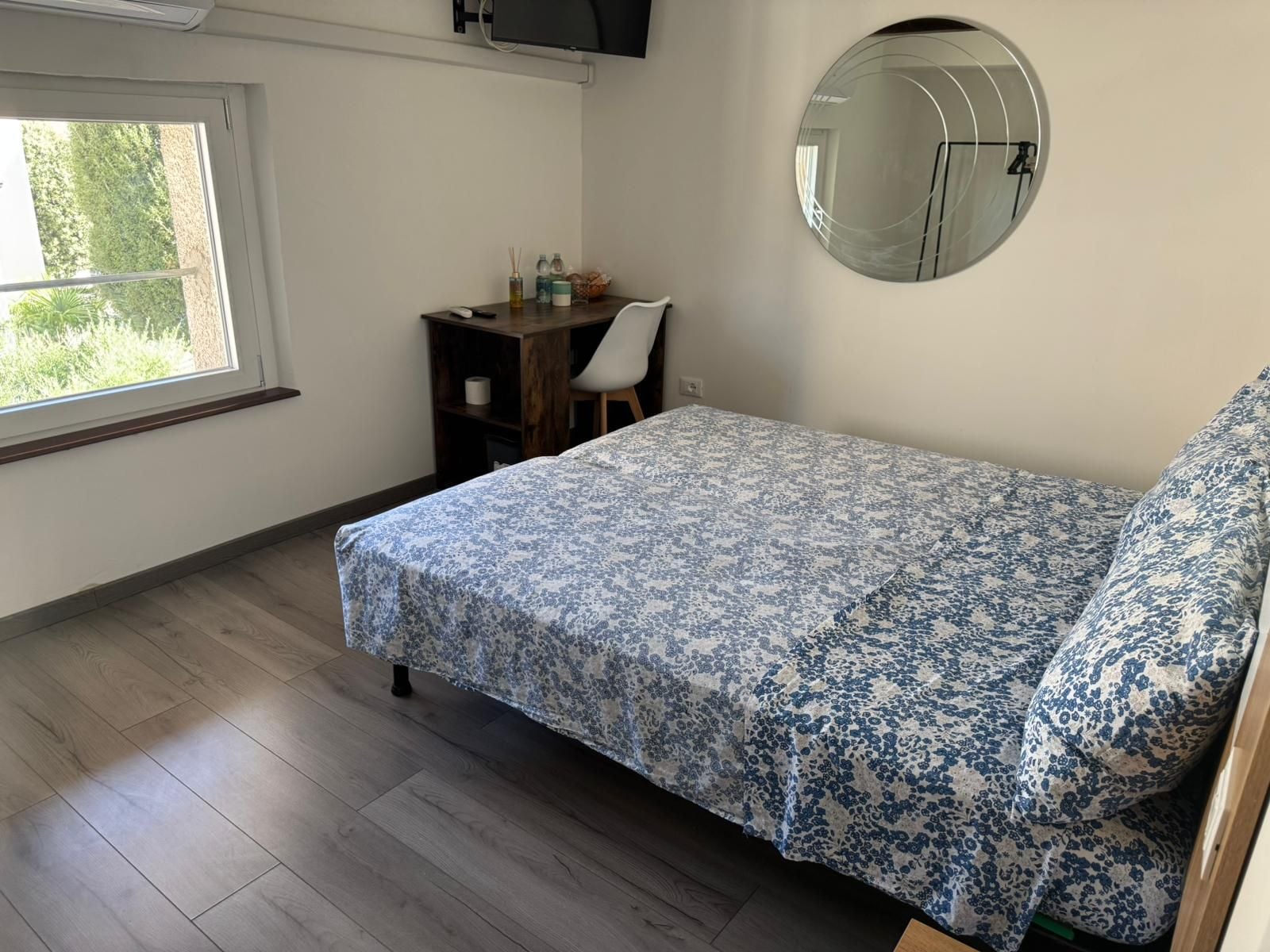 Une chambre à coucher avec un lit, un bureau, une chaise, un miroir rond et une fenêtre.