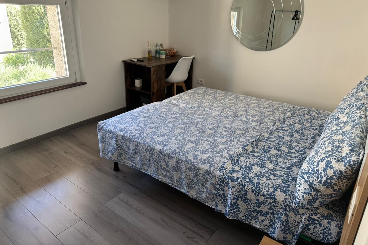 A bedroom with a bed, a desk, a chair, a round mirror, and a window.