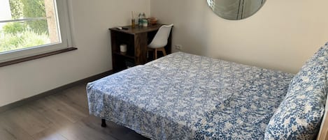 A bedroom with a bed, a desk, a chair, a round mirror, and a window.