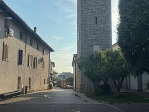 Una calle estrecha bordeada de edificios, uno con una torre de piedra, y una acera arbolada.