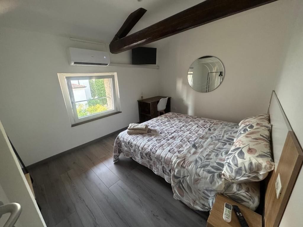 A bedroom with a bed, a bedside table, a mirror, a window, and an air conditioning unit.