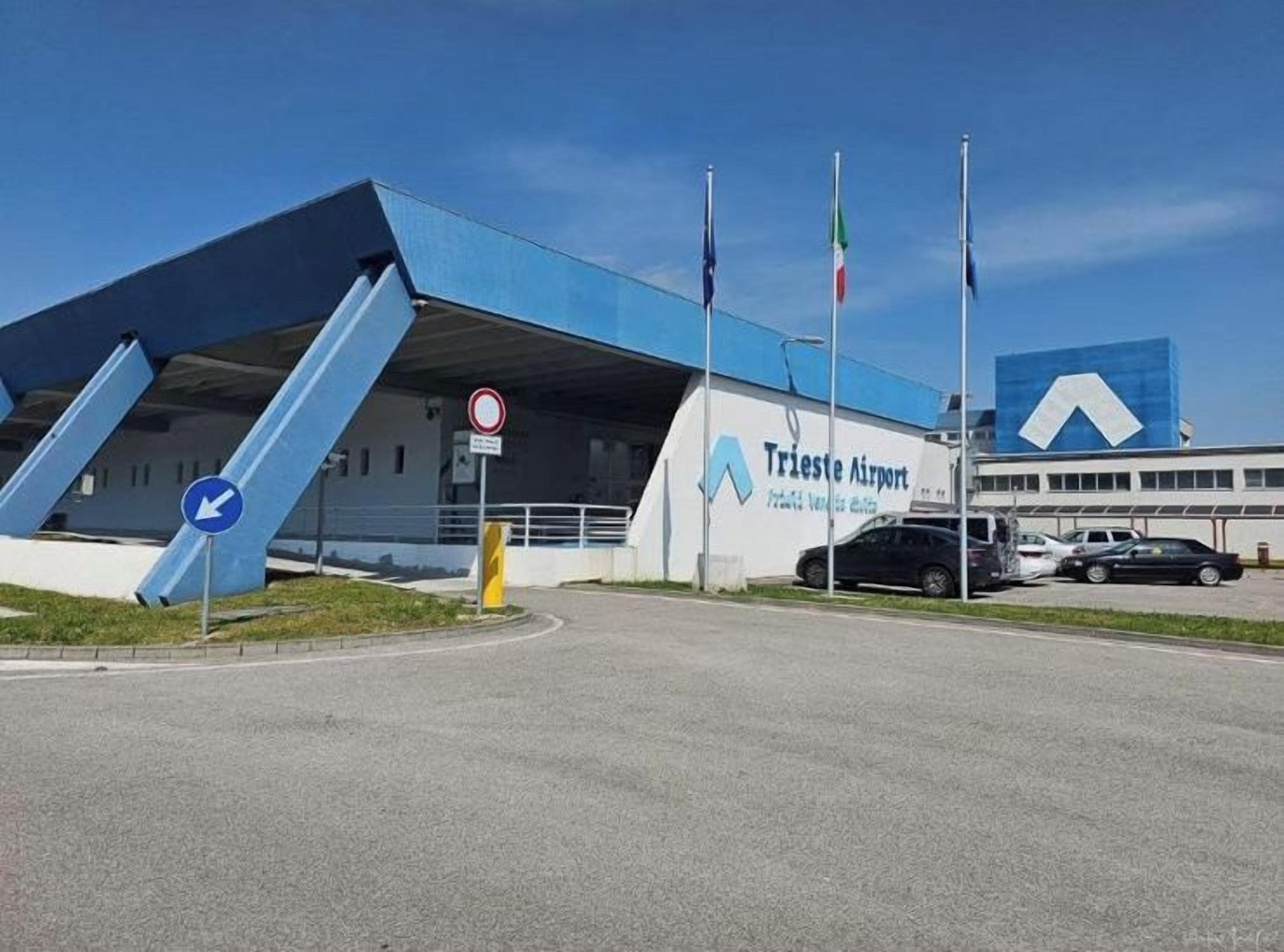 An airport terminal with a blue roof, a sign for Trieste Airport, and a no parking sign.