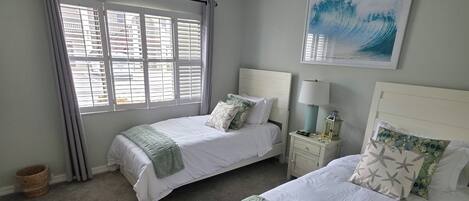 A bedroom with two beds, a ceiling fan, a window with blinds, a wall-mounted painting, and a bedside table with a lamp.