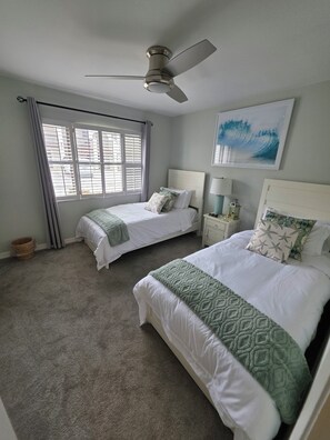 Ein Schlafzimmer mit zwei Betten, einem Deckenventilator, einem Fenster mit Jalousien, einem Wandbild und einem Nachttisch mit Lampe.