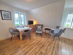 A room with a wooden floor, a dining table with chairs, a TV, a lamp, and a window with curtains. - Tewelken Apartment - Tewelken Apartment in Wrixum - quiet location, modern (Wrixum)