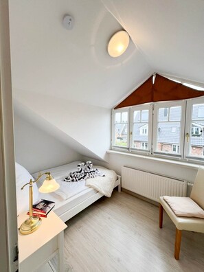 A small, well-lit attic bedroom with a single bed, a chair, a desk with a lamp, and a window overlooking a residential area. - Leyla OG - Leyla Vacation Rental on Sylt: Balcony, Parking, and Dog-Friendly (Sylt)