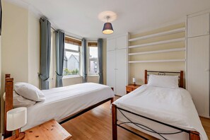 A bedroom with two single beds, a bedside table with a lamp, a wardrobe, and a window with curtains. - Host & Stay | House On The Hill (Hastings)
