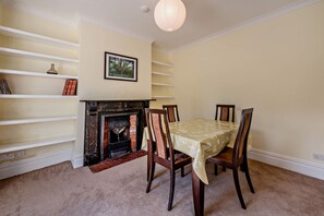 A dining room with a fireplace, wooden chairs, a table with a tablecloth, and a wall-mounted shelf with books and decorative items. - Host & Stay | House On The Hill (Hastings)