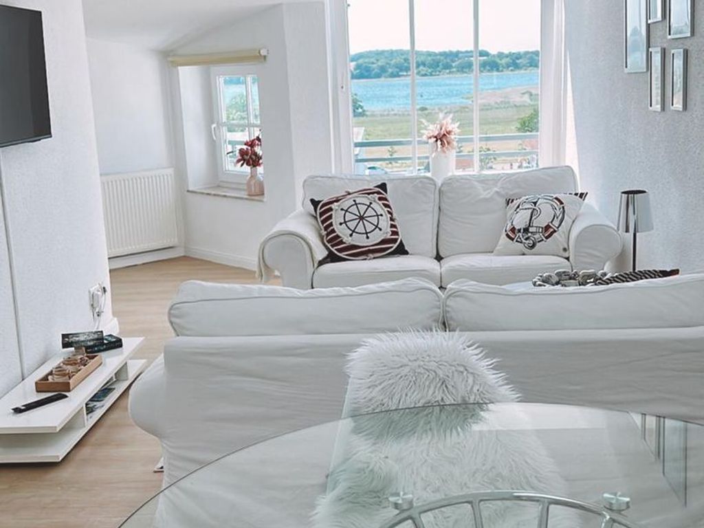 Een moderne woonkamer met een witte zithoek, een glazen salontafel en uitzicht op zee door grote ramen.