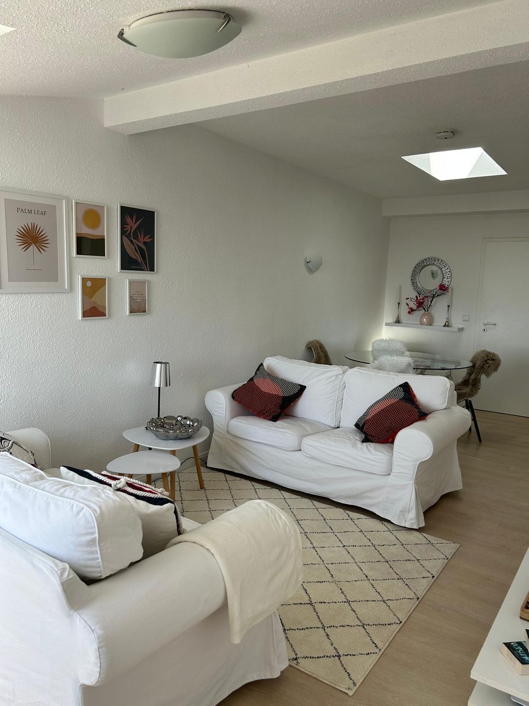Ein modernes Wohnzimmer mit einem weißen Sofa, einem kleinen runden Tisch und einem Couchtisch. An der Wand befinden sich gerahmte Kunstwerke und ein dekorativer Spiegel über dem Sofa.