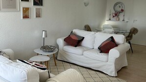 Ein modernes Wohnzimmer mit einem weißen Sofa, einem kleinen runden Tisch und einem Couchtisch. An der Wand befinden sich gerahmte Kunstwerke und ein dekorativer Spiegel über dem Sofa.