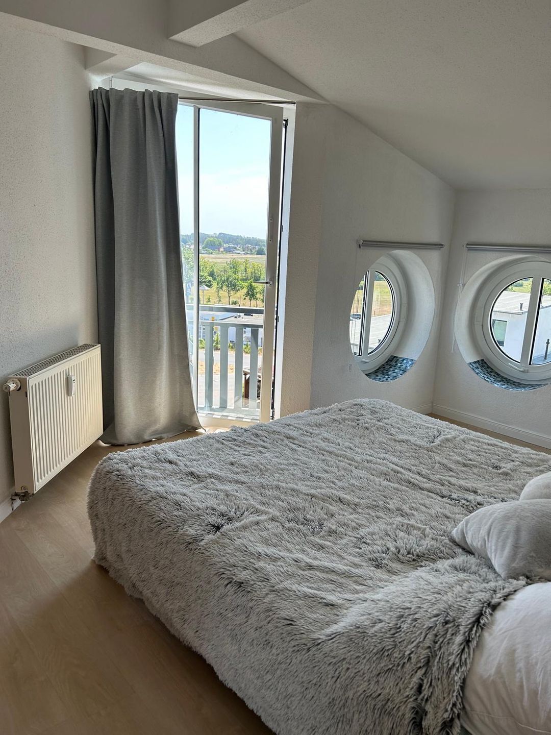 Ein Schlafzimmer mit einem großen Bett, einem Fenster mit Vorhängen, einem Heizkörper und Blick ins Freie.