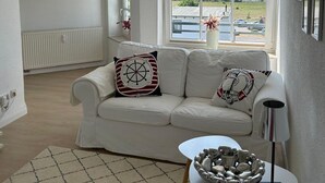 Ein Wohnzimmer mit einem weißen Sofa, einem kleinen Tisch mit einer dekorativen Schale und einem großen Fenster mit Blick auf einen See und viel Grün.