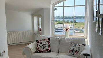 Ein Wohnzimmer mit einem weißen Sofa, einem kleinen Tisch mit einer dekorativen Schale und einem großen Fenster mit Blick auf einen See und viel Grün.