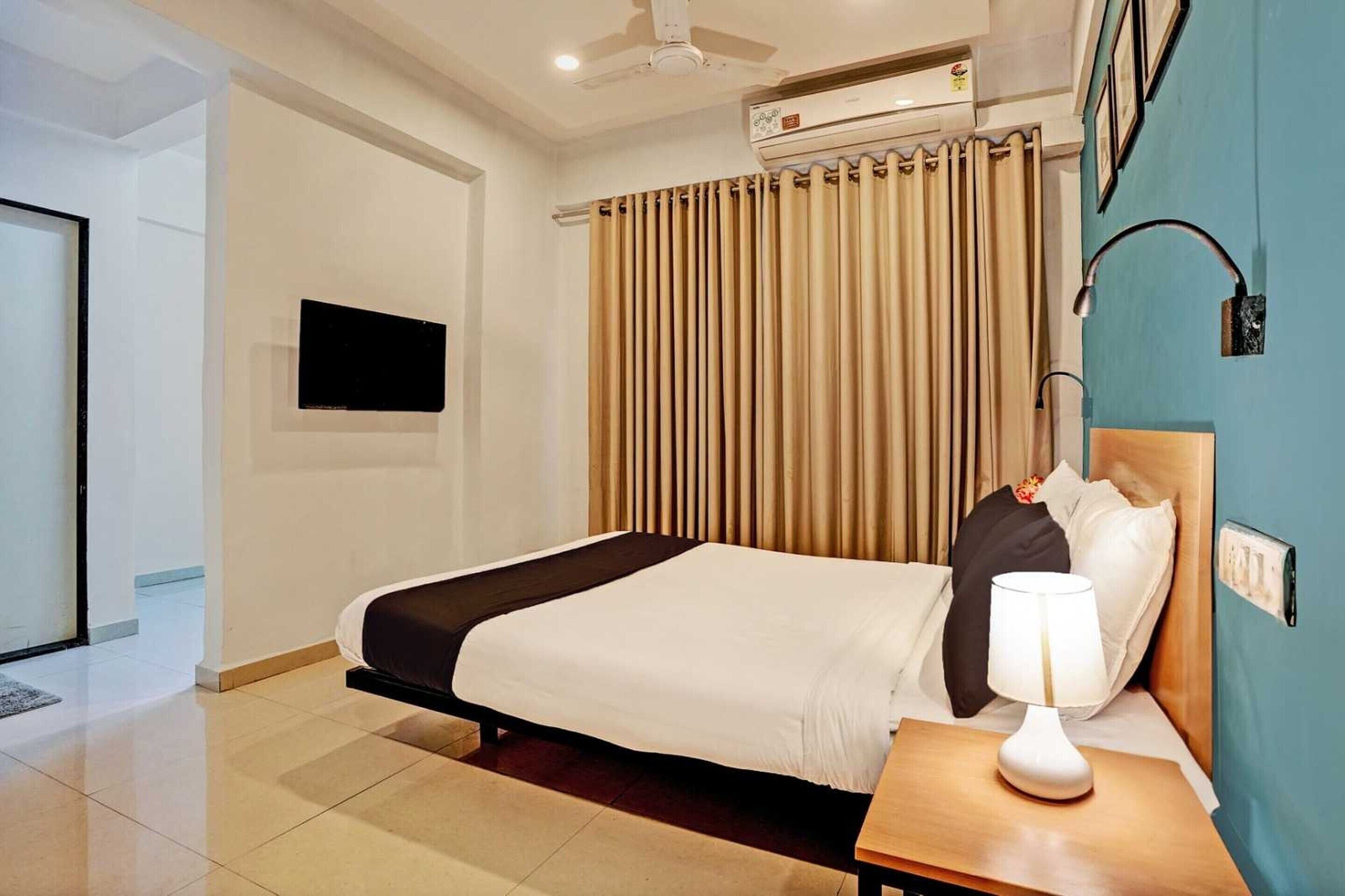 A hotel room with a bed, a bedside table with a lamp, a wall-mounted TV, and a window with curtains.