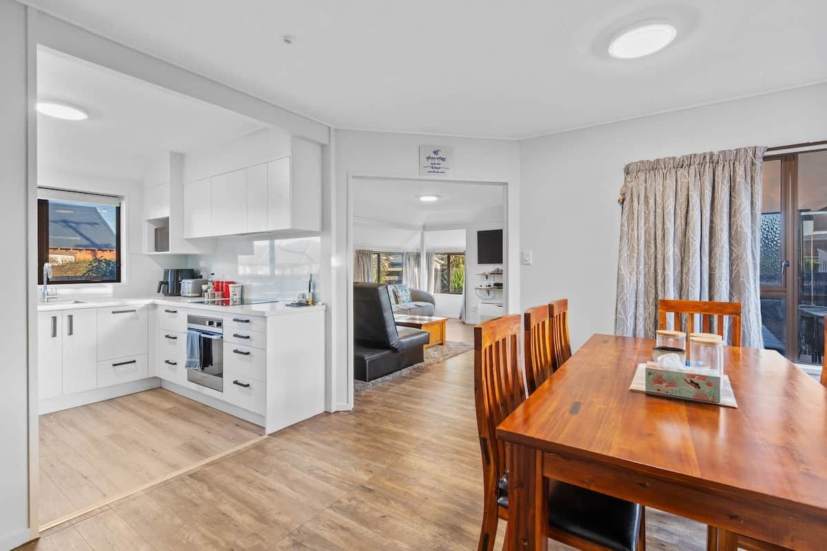 A modern kitchen with white cabinetry, a dining area with a wooden table, and a living room with a sofa and a TV.