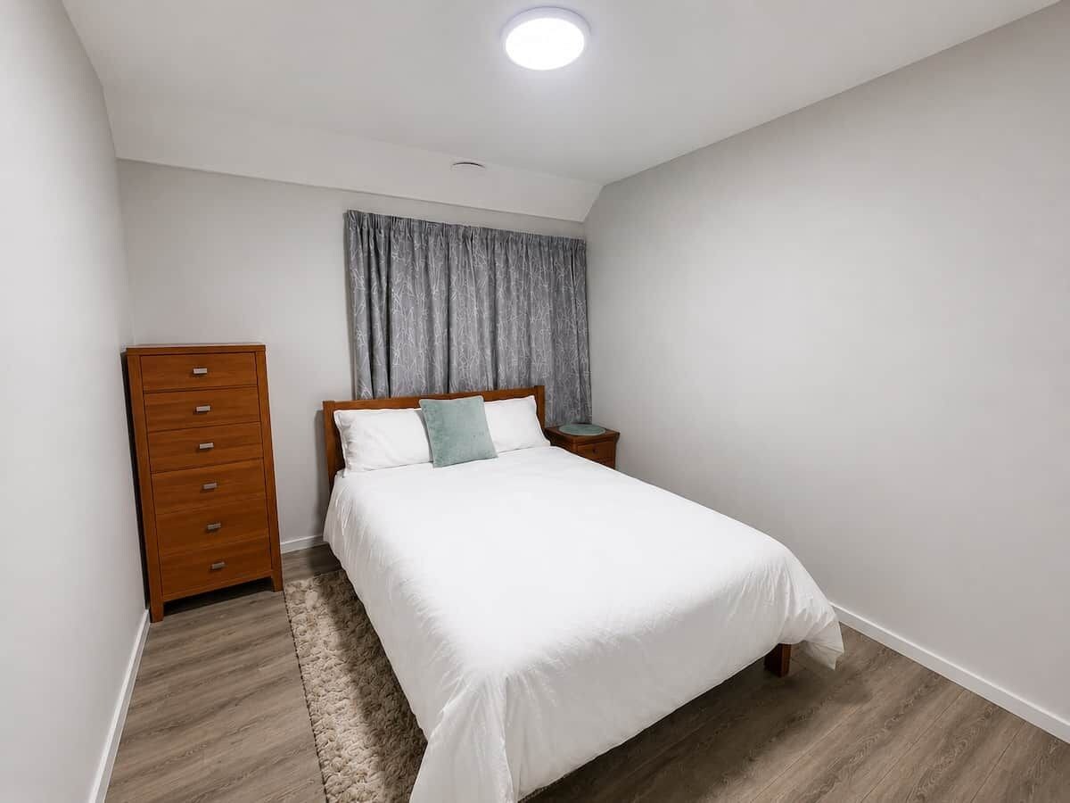 Quarto com cama, penteadeira de madeira, mesa de cabeceira e janela com cortinas.