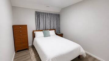 Quarto com cama, penteadeira de madeira, mesa de cabeceira e janela com cortinas.