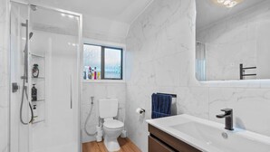 Une salle de bain moderne avec une douche à l’italienne en verre, des toilettes blanches, une grande baignoire et une fenêtre laissant entrer la lumière naturelle.