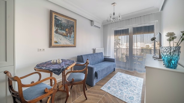 A living room with a sofa, a dining table with chairs, a painting on the wall, and a view through the window. - Kierbedzia Elegant Classic Blinds, Free Parking, Mokotow, Warsaw (Warszawa)